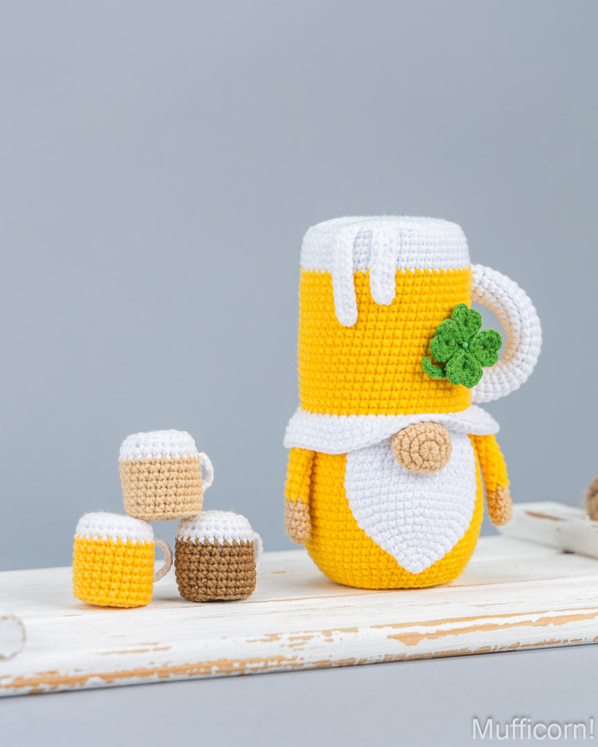 Crocheted beer mug gnome figurine in yellow and white with a green shamrock accent, standing on a rustic white wooden surface next to three small crocheted beer mugs against a solid gray background.