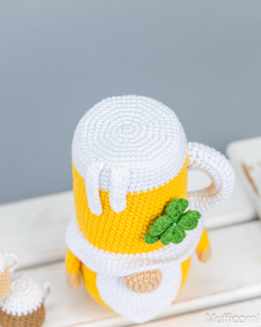 A crocheted beer mug gnome wearing a white foam-topped mug cover with a green shamrock accent, standing on a white wooden surface against a soft gray background.