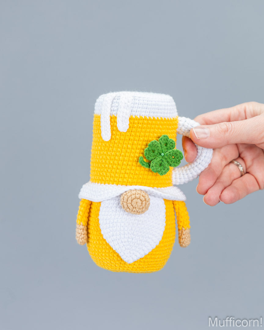 A hand holds a crocheted gnome mug cozy in yellow and white with a green shamrock detail, against a solid gray background.