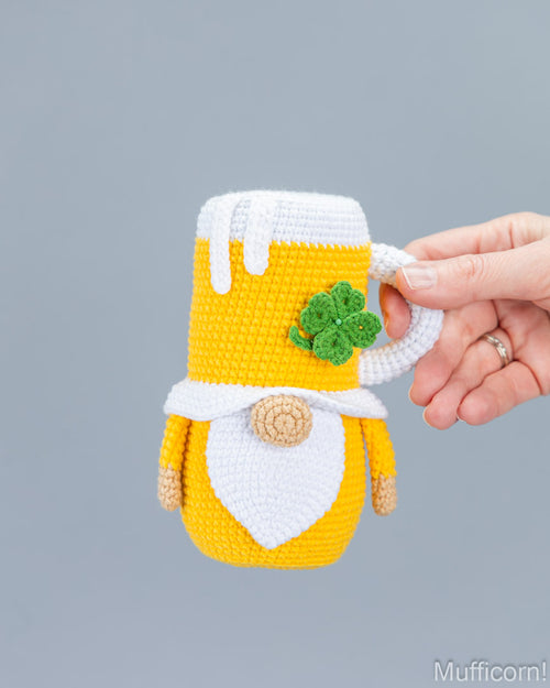 A hand holds a crocheted gnome mug cozy in yellow and white with a green shamrock detail, against a solid gray background.