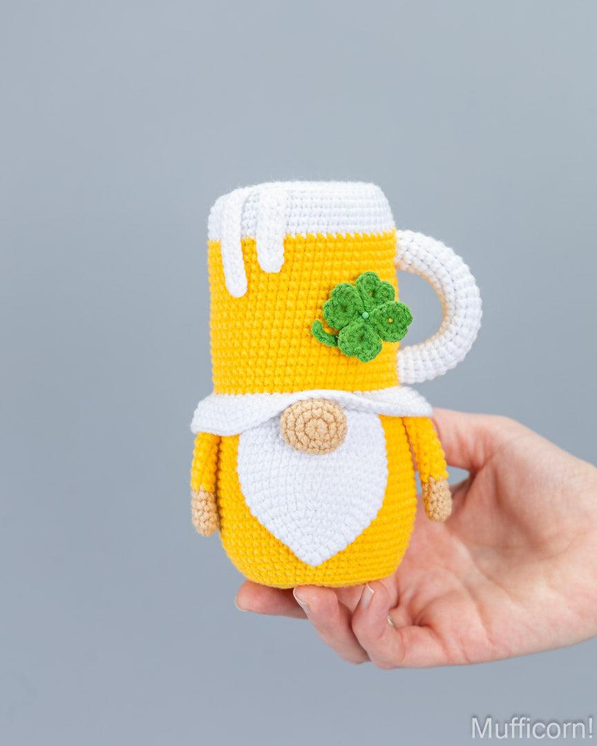 A hand holds a crocheted St. Patrick's Day mug gnome in yellow, white, and green.