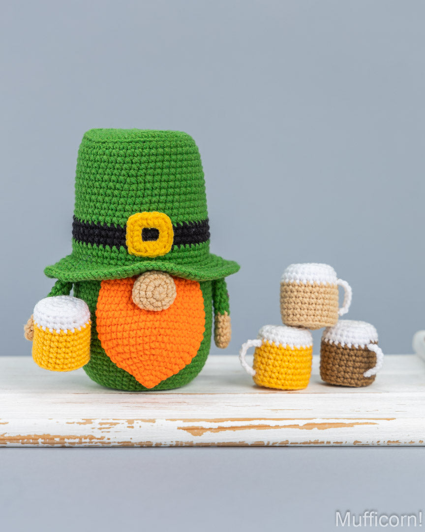 Crocheted leprechaun doll wearing a green hat with a black band and yellow buckle, an orange beard, and holding small beer mugs, displayed on a rustic white table against a light gray background. Three additional beer mugs sit to the right.