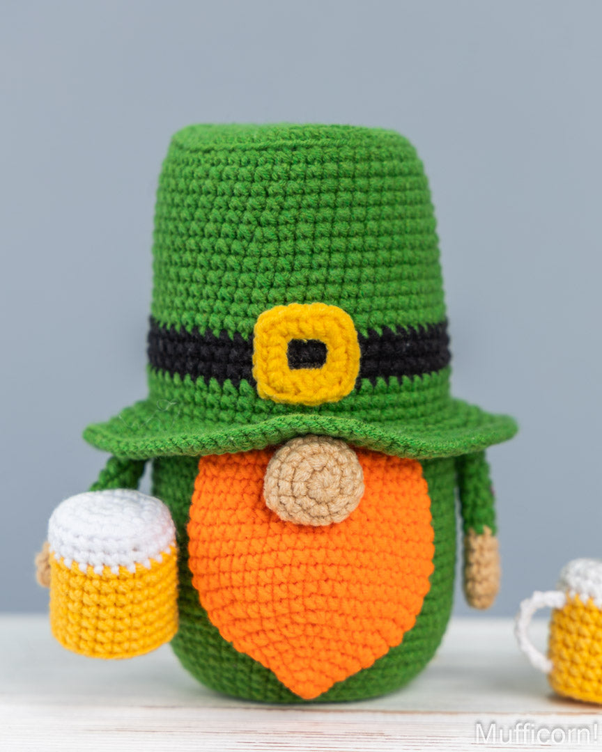 A crocheted leprechaunt figure wearing a green hat with a black band and yellow buckle, an orange beard, and holding two small beer mugs, shown against a soft gray background.
