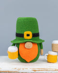 Crocheted St. Patrick's Day gnome figurine wearing a green hat with a black band and yellow buckle, orange beard, and holding two yellow and white beer mugs, displayed on a rustic white wooden surface against a gray background.