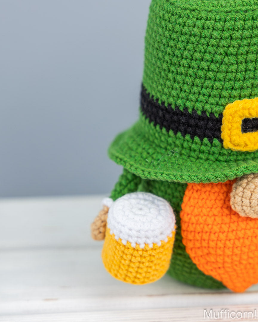Close-up of a crocheted leprechaun figure wearing a green hat with a black band and yellow buckle, holding a small yellow mug with white foam, on a light wooden surface against a soft gray background.