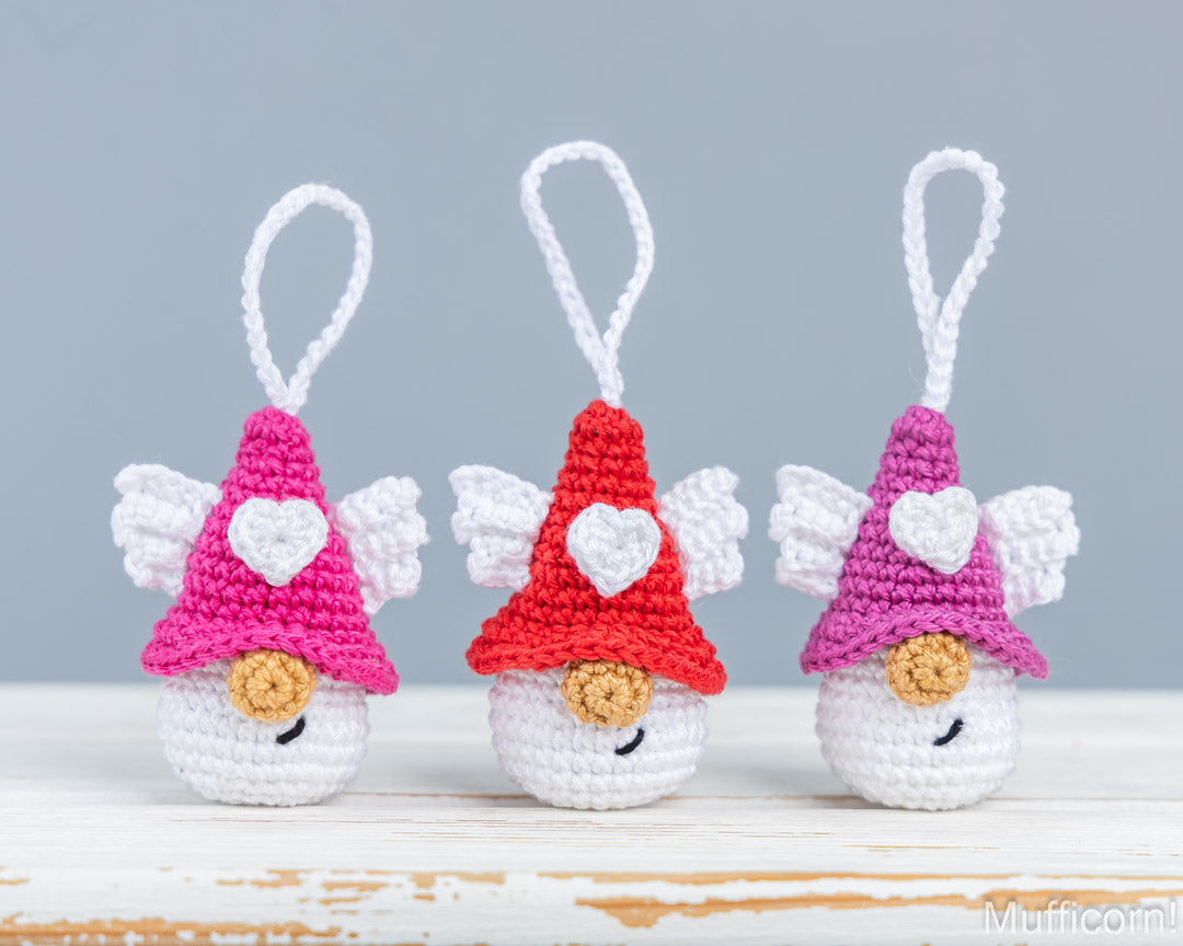 Three crocheted angel ornaments in pink, red, and purple, each with white wings, a white heart on their hats, and white hanging loops, displayed on a white wooden surface against a gray background.