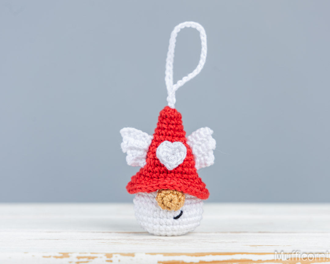 A small crocheted angel ornament with a red hat, white wings, and a white loop at the top, photographed on a light surface against a soft gray background.