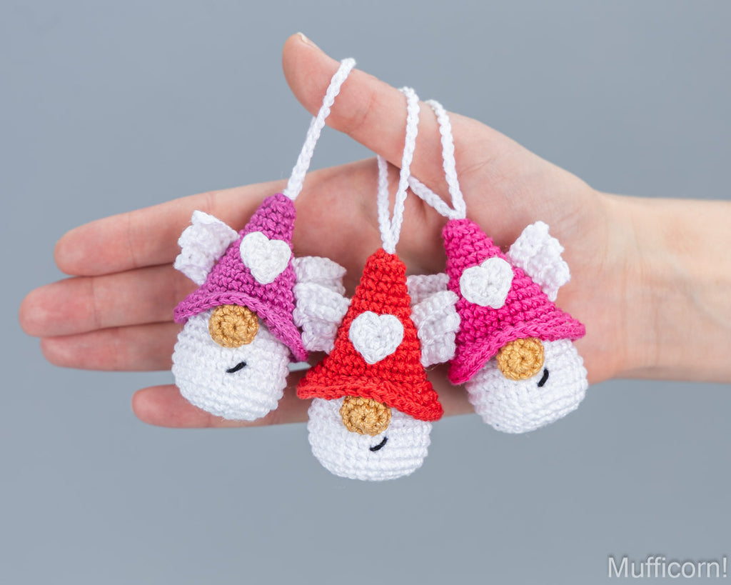 Three small crocheted angel ornaments are held in a person's hand against a plain gray background. Each ornament has white wings, a white face, and a pointed hat in pink, purple, or red, with a white heart on the hat and a small gold circular detail below the nose.