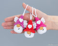 Three small crocheted angel ornaments are held in a person's hand against a plain gray background. Each ornament has white wings, a white face, and a pointed hat in pink, purple, or red, with a white heart on the hat and a small gold circular detail below the nose.