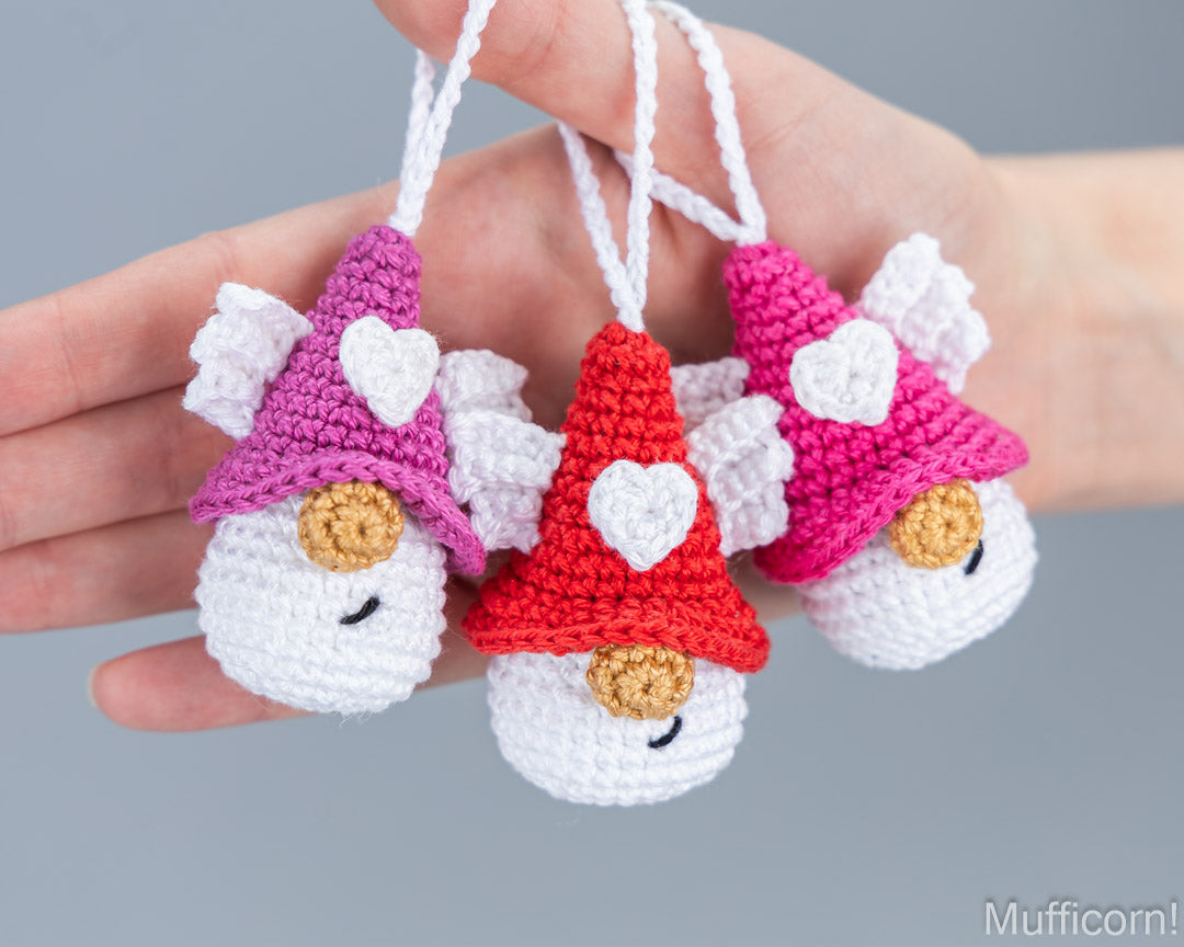 Three small crocheted mufficorn ornaments are held in a person's hand against a plain gray background. Each ornament has a white body, a pointed hat in pink, red, or purple, a white heart on the hat, and a small tan nose.