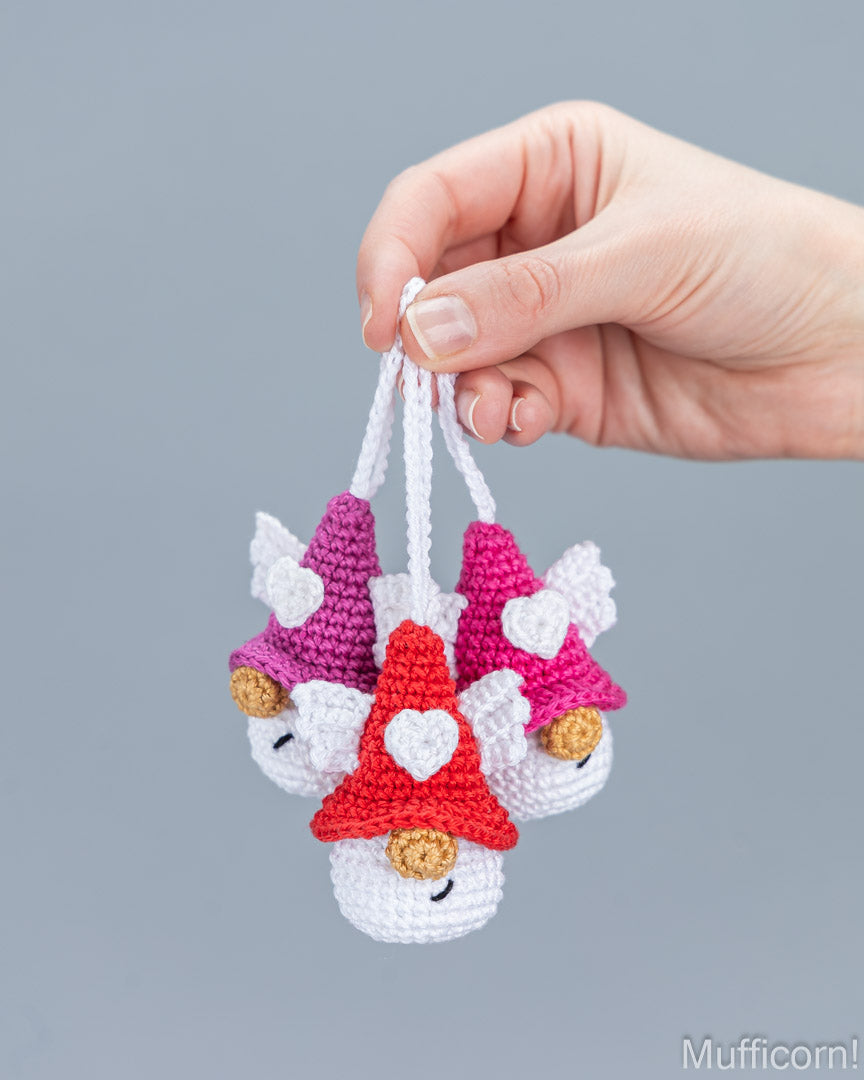 A hand holding three small crocheted gnome ornaments with white wings and colorful hats in red, pink, and purple against a solid gray background.