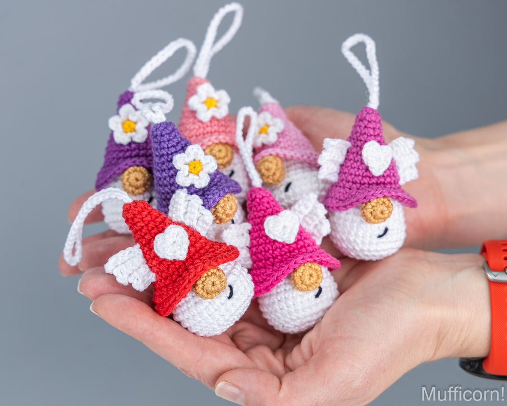 Seven small crocheted gnome ornaments in red, pink, purple, and peach are held in a person's hand against a plain gray background. Each ornament has a pointed hat, white wings, a flower on the hat, and a white hanging loop.