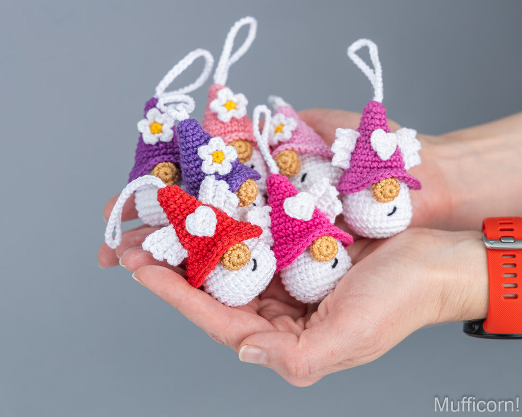 Five small crocheted gnome ornaments in red, pink, purple, and peach hang from white loops and are held in a person's hands against a plain gray background.