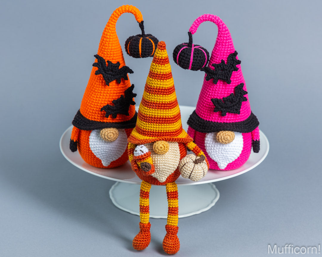 Three crocheted Halloween gnome dolls with pointed hats and pumpkin accents, displayed on a white cake stand against a gray studio background. The gnomes are orange, yellow, and pink, with black bats and hats and white beards.