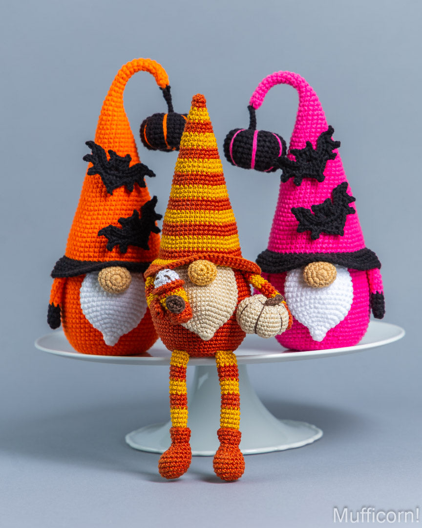 Three crocheted Halloween gnome dolls displayed on a white cake stand against a gray studio background. The central gnome wears a striped orange and yellow hat and holds a small pumpkin.