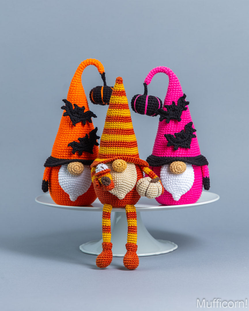 Three crocheted Halloween gnomes in orange, yellow, and pink hats sit on a white pedestal against a gray background. The gnomes have white beards, brown buttons, and black bat accents on their hats.