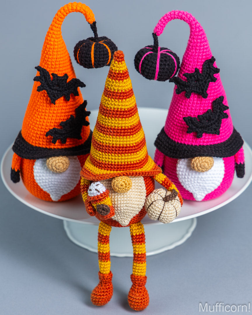 Three crocheted Halloween gnome decorations displayed on a white pedestal against a light gray background. The gnomes have pointed hats with black bat appliqués and small pumpkin accents, with one orange, one yellow-and-orange striped, and one bright pink.