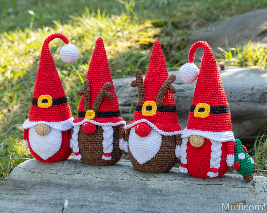Four crocheted Christmas gnomes with red pointed hats, white beards, and brown bodies, arranged outdoors on a log with grass in the background. Two gnomes have reindeer antlers and one has a small green tree accessory.