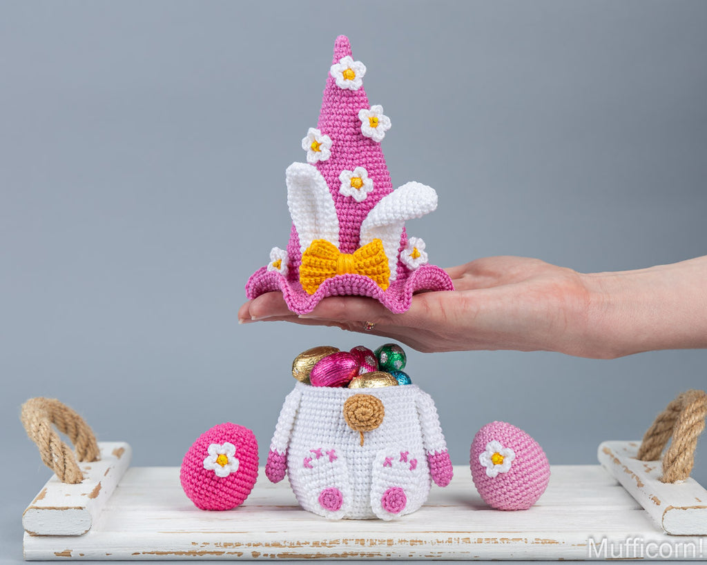 A crocheted Easter-themed decoration set displayed on a white wooden surface against a gray background. A human hand holds a pink and white crocheted bunny hat with a yellow bow and white flowers.