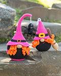 Two crocheted witch gnomes with pink and black hats, orange yarn beards, and one hanging black spider decoration, displayed outdoors on a stone surface.