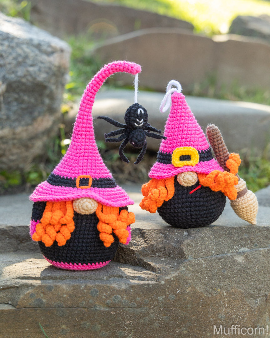 Two crocheted witch gnomes with pink and black hats, orange yarn beards, and one hanging black spider decoration, displayed outdoors on a stone surface.