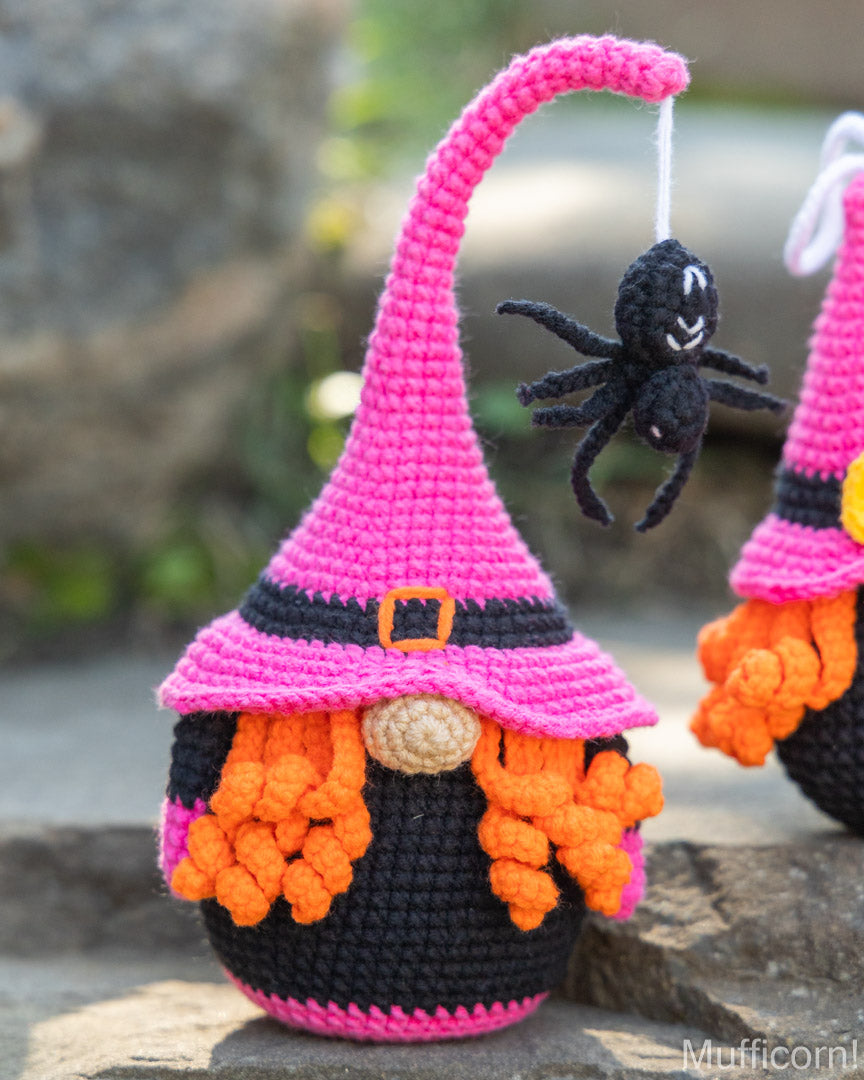 A crocheted witch gnome decoration with a bright pink pointed hat, black body, orange yarn hair, and a small black spider hanging from the hat, photographed outdoors on a stone surface.