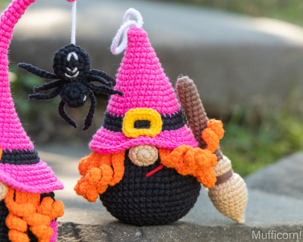 A crocheted witch doll with a pink hat, black body, orange yarn hair, and a broomstick, photographed outdoors on a stone surface with a black spider hanging nearby.