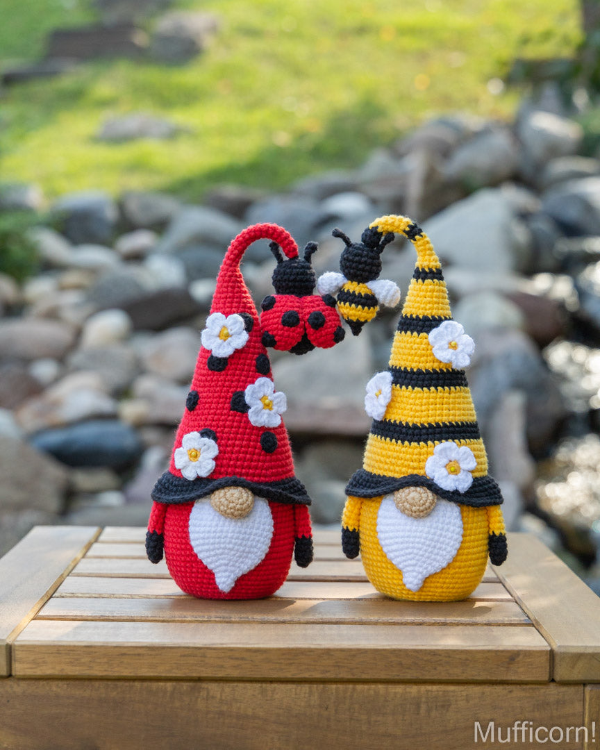Two crocheted gnome garden figures posed on a wooden table outdoors, with a blurred rocky garden background. One gnome is red with black spots and a black hat, the other is yellow and black striped with a bee figure on its hat, both decorated with white flowers.