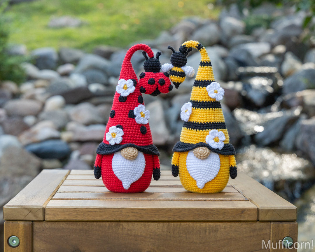 Bee gnome and ladybug gnome  posed on a wooden crate outdoors, with a blurred rocky stream and grass in the background. The left gnome is red with black and white details, and the right gnome is yellow and black with white flower accents.