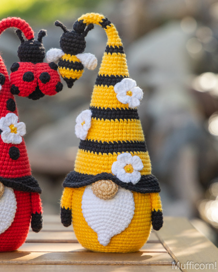 A crocheted bumblebee gnome doll with a tall yellow and black striped hat, white flower accents, and a white beard, standing on a wooden surface with a blurred outdoor background. Part of a red ladybug gnome is visible on the left.