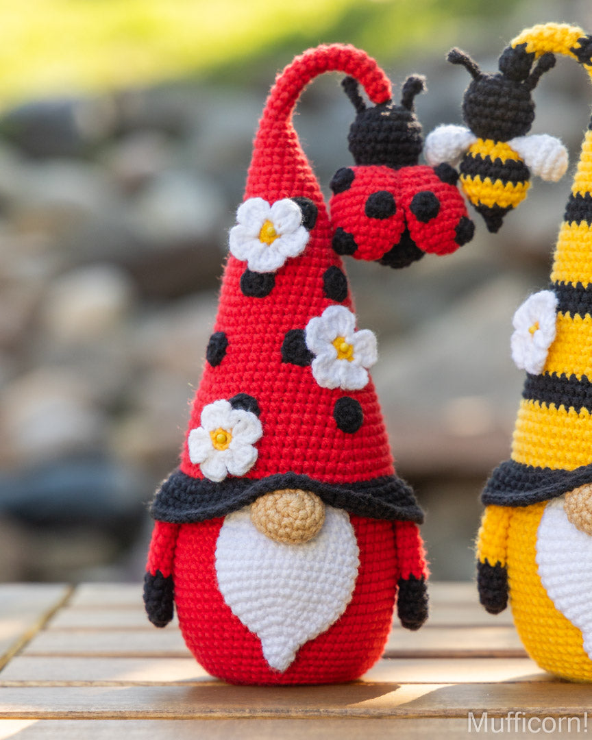 A crocheted ladybug gnomel in red with black spots, white flowers, and a ladybug on its hat, standing on a wooden surface outdoors. Part of a yellow-and-black striped gnome is visible at the right edge.