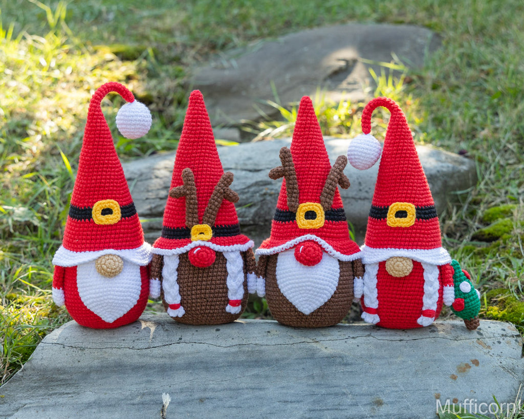 Four crocheted Christmas gnome dolls standing side by side on a weathered log outdoors, with grass and rocks in the background. The gnomes are red and white with black, yellow, brown, and gold details, including two with reindeer antlers.