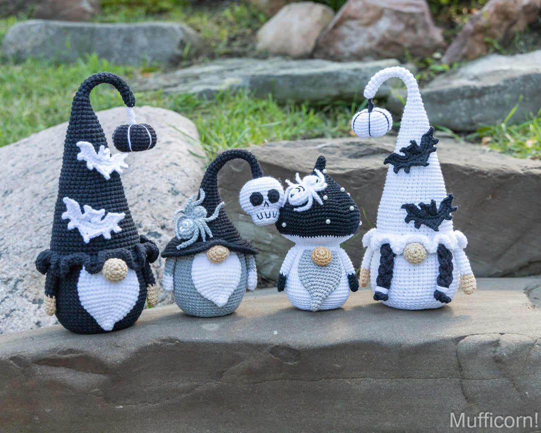 Four crocheted Halloween gnomes in black, white, and gray are arranged on a stone surface outdoors with grass and rocks in the background.
