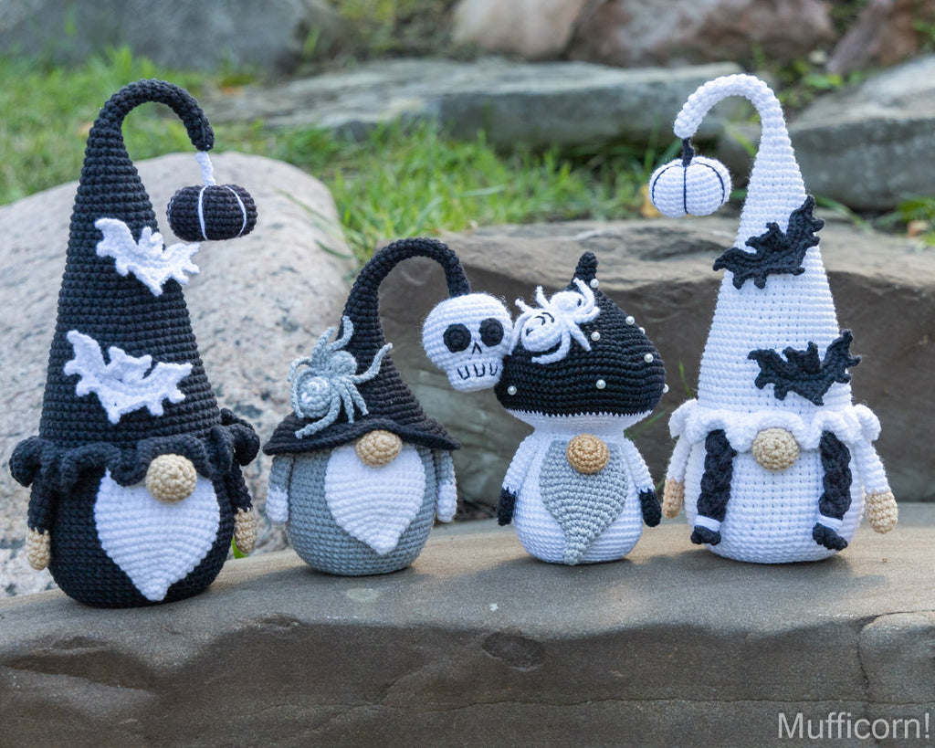 Four crocheted Halloween gnomes arranged outdoors on a stone surface with grass and rocks in the background. The gnomes are black and white with spiderwebs, bats, pumpkins, and one has a skull on its hat.