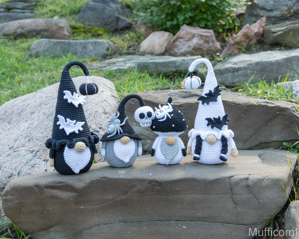 Four crocheted Halloween gnomes in black and white outfits posed on a large stone outdoors, with grass, rocks, and plants in the background.