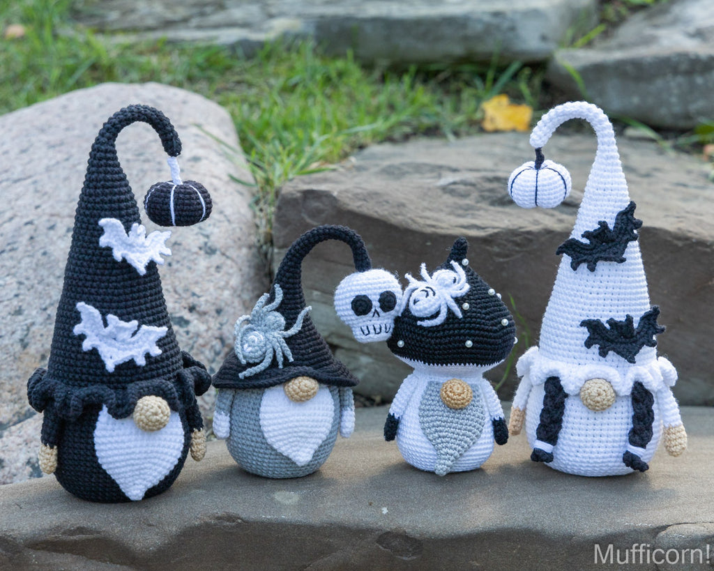 Four crocheted Halloween gnomes with pointed hats, arranged outdoors on a stone surface with grass and rocks in the background. The gnomes are black and white with small decorative details like bats, spiders, skulls, and pumpkins.
