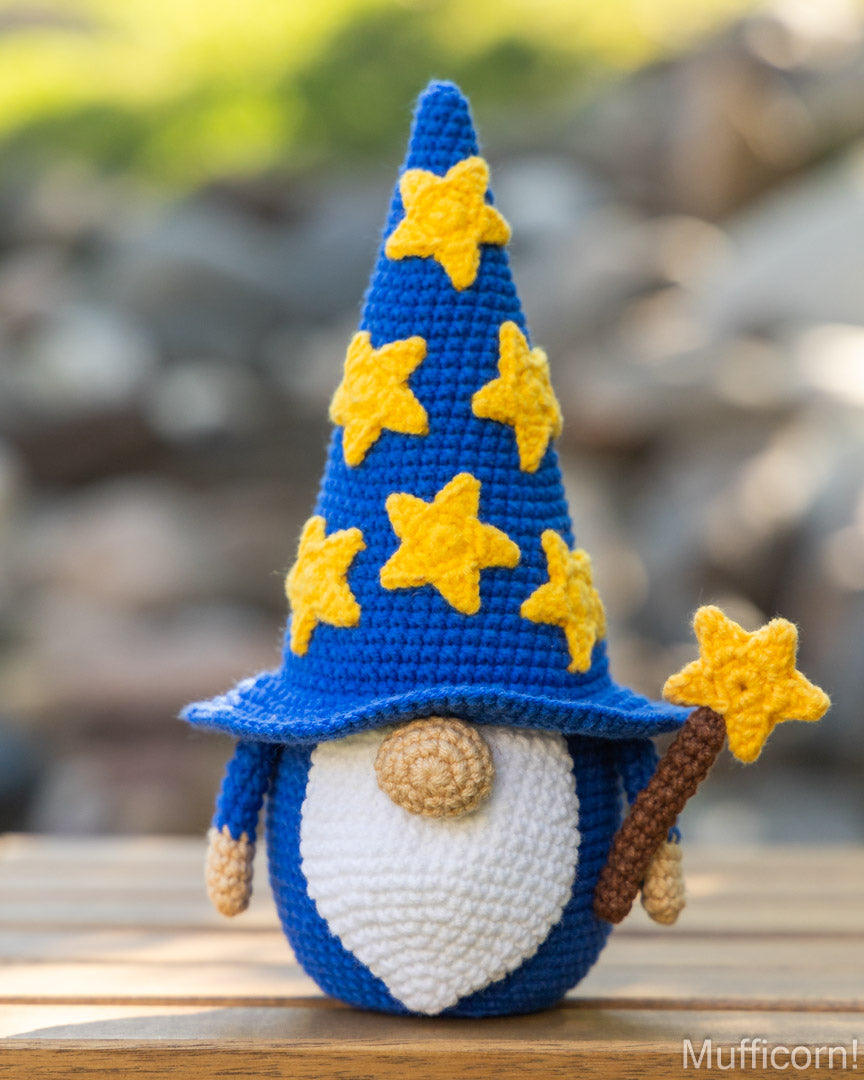 A crocheted wizard gnomem with a tall blue hat decorated with yellow stars, a white belly, and a brown staff, standing on a wooden surface with a blurred outdoor background.