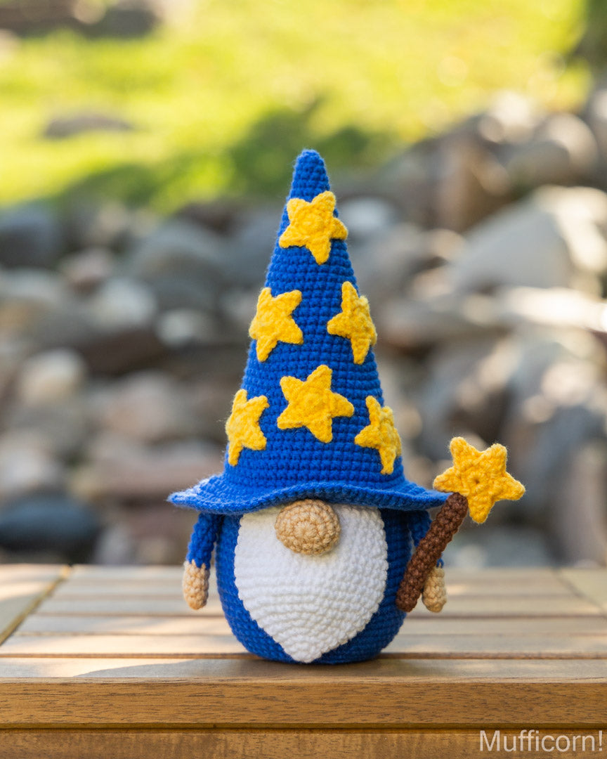 A crocheted wizard gnome sits on a wooden surface outdoors with a blurred background of rocks and greenery. The gnome has a tall blue hat with yellow star details, a white beard, and holds a small yellow star wand.