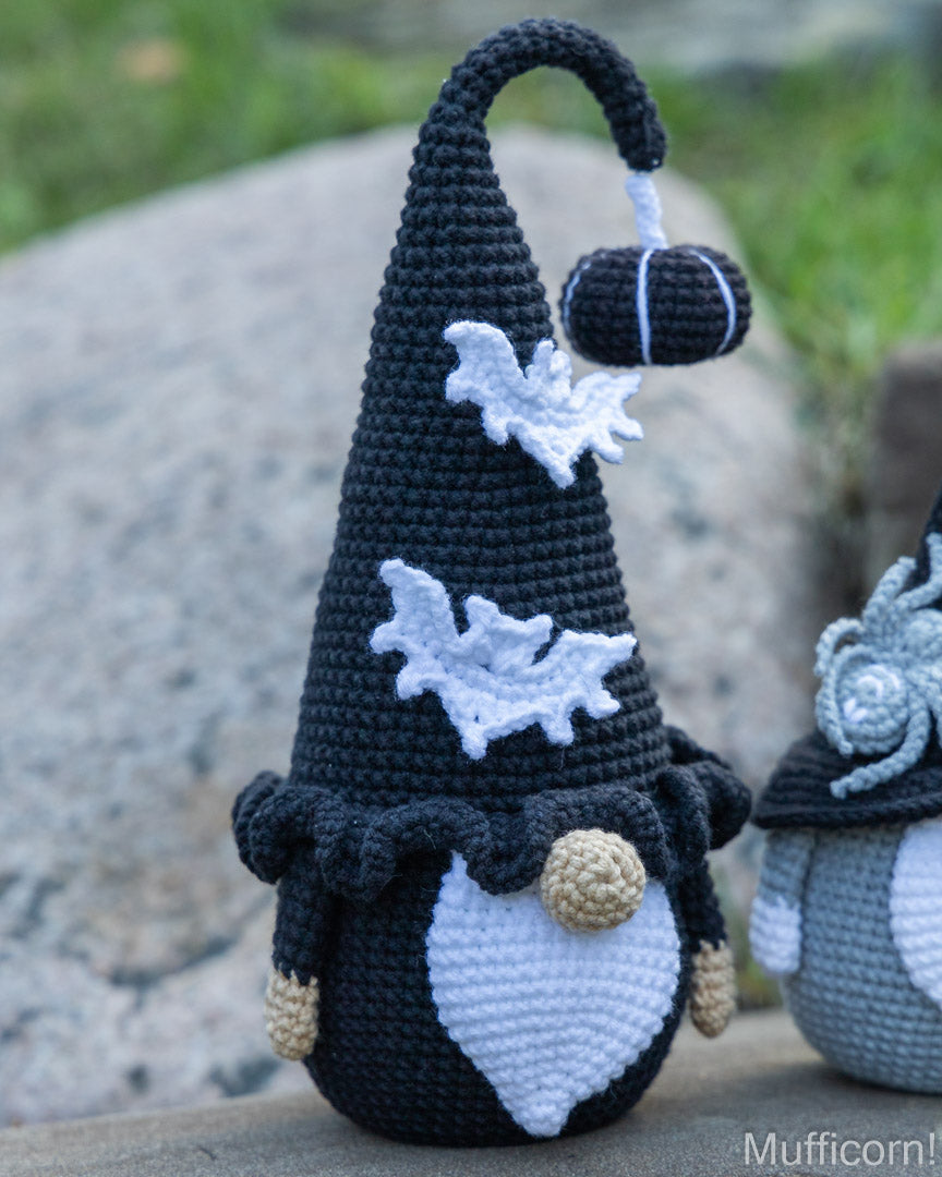 A crocheted black gnome doll with a tall pointed hat, white bat decorations, a white beard, and tan hands, photographed outdoors on a stone surface with a blurred grassy background. A second similar crocheted gnome is partially visible on the right.