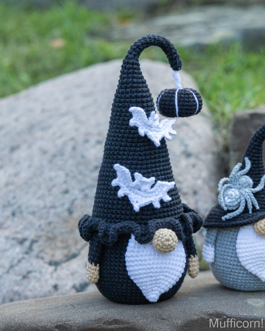 Handmade crocheted Halloween gnome doll with a tall black pointed hat, white bat details, and a white beard, standing on a stone outdoors with another gnome partially visible.
