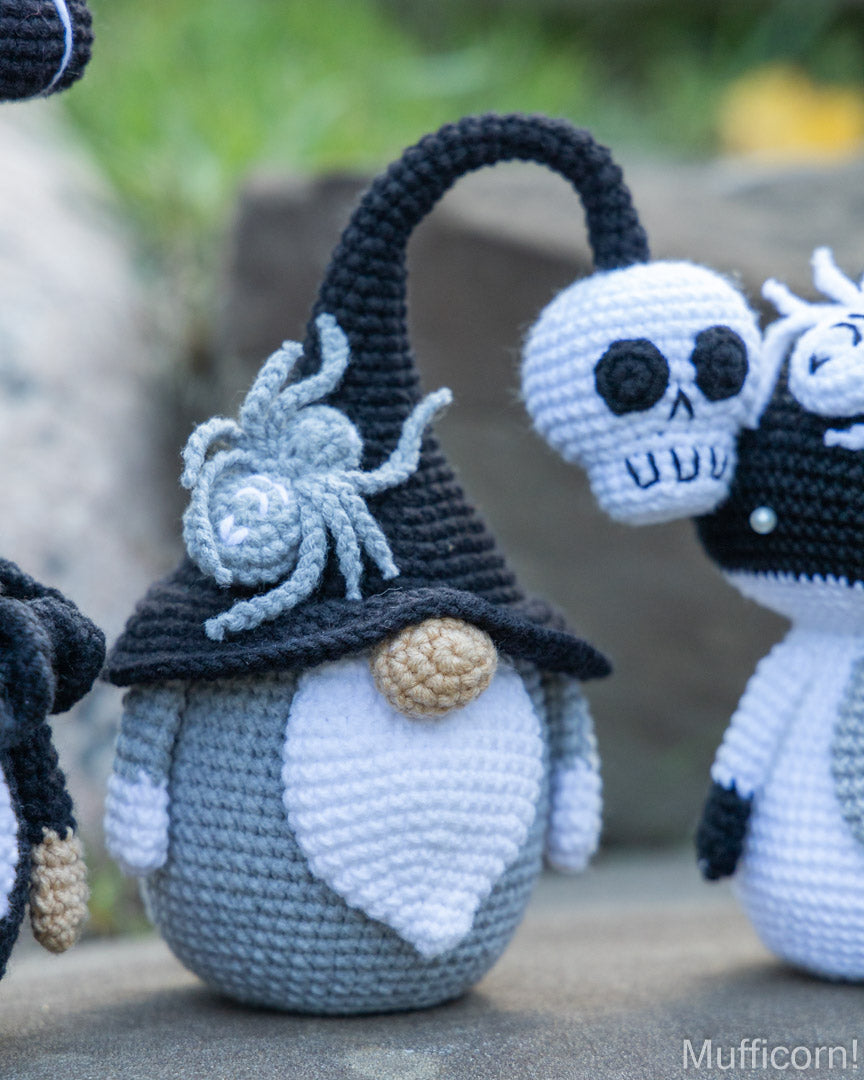 A crocheted Halloween gnomemuffler doll with a tall black hat, white beard, and a gray spider on the brim, shown outdoors next to other crocheted gnomes. The brand name 'Mufficorn!' is visible in the bottom right corner.
