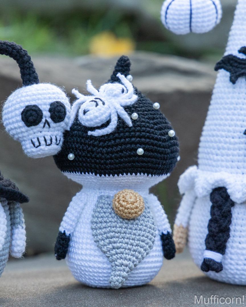 A crocheted muffin character toy in black and white with a white spider on its head and a small skull accessory, shown outdoors with other similar crocheted figures partially visible.