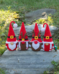 Four crocheted Christmas gnomes in holiday outfits arranged on a gray stone slab outdoors, with grass and rocks in the background.
