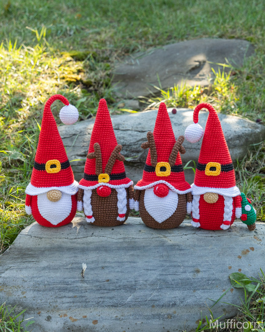 Four crocheted Christmas gnomes in holiday outfits arranged on a gray stone slab outdoors, with grass and rocks in the background.