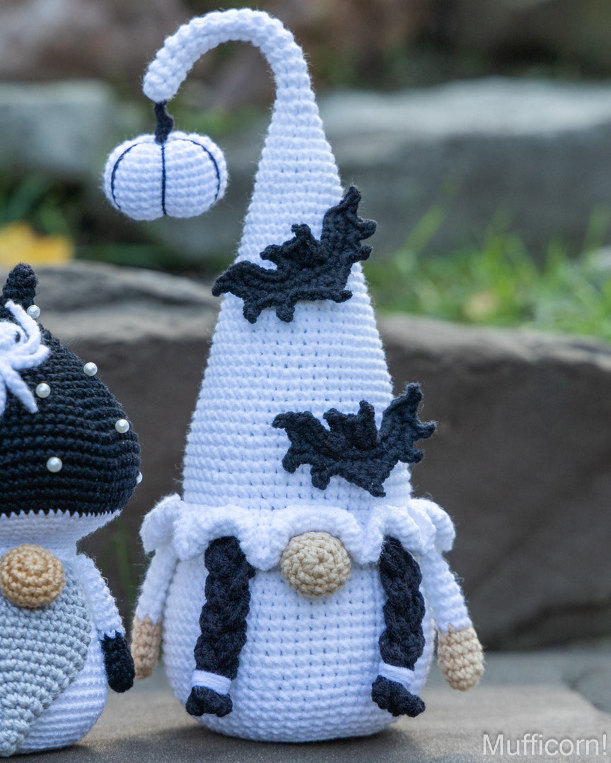 A crocheted Halloween gnome doll with a tall white pointed hat, black bat details, and a small hanging pumpkin, shown outdoors on a stone surface with another crocheted gnome partially visible at the left edge.