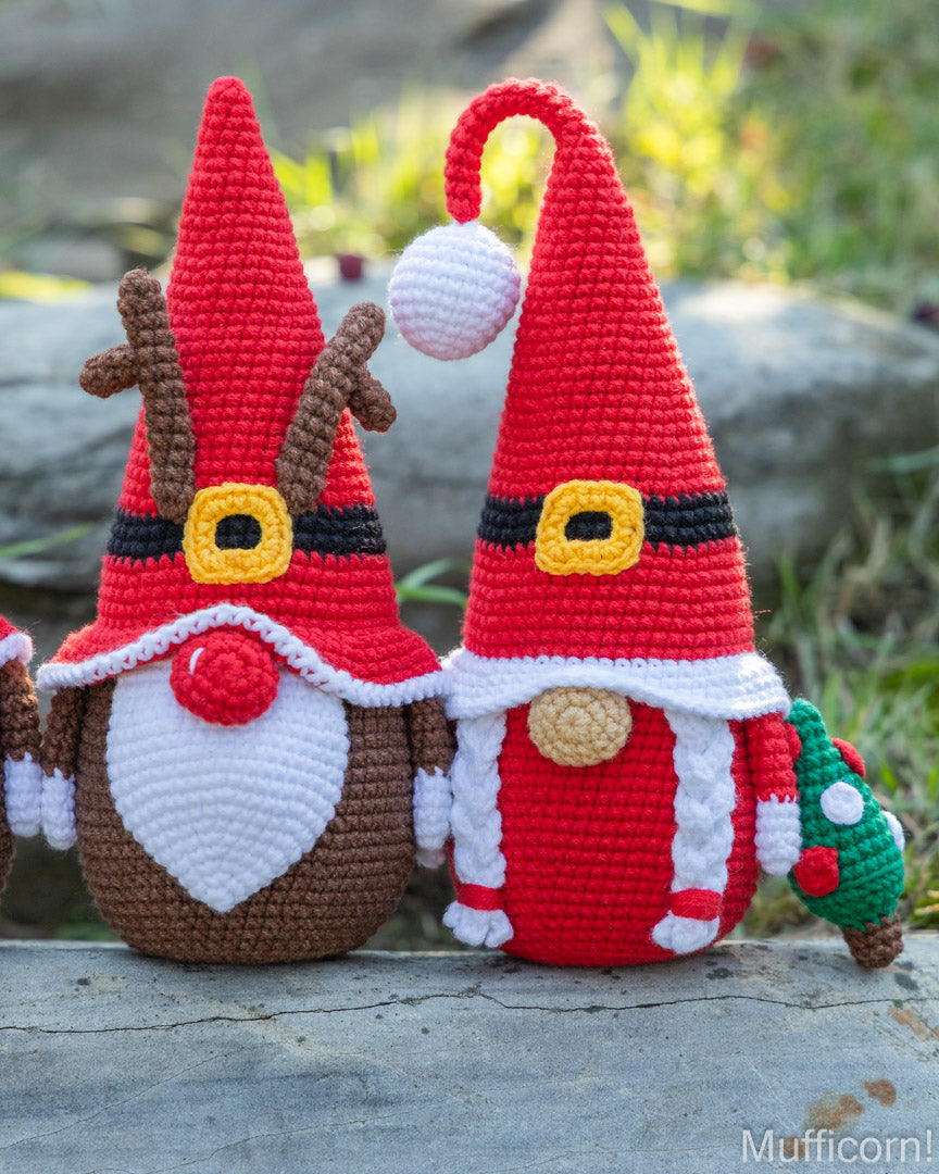 Two crocheted Christmas gnomes posed outdoors on a stone surface with grass and rocks blurred in the background. One gnome has brown antlers and a white beard, while the other wears a red hat with a white pom-pom and holds a small green tree.