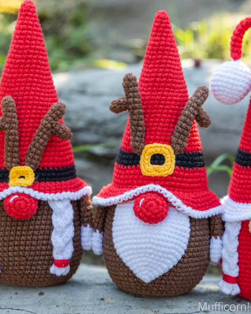 Three crocheted Christmas gnome ornaments with red pointed hats, brown bodies, white beards, and black-and-yellow belts, photographed outdoors on a stone surface with blurred greenery in the background.