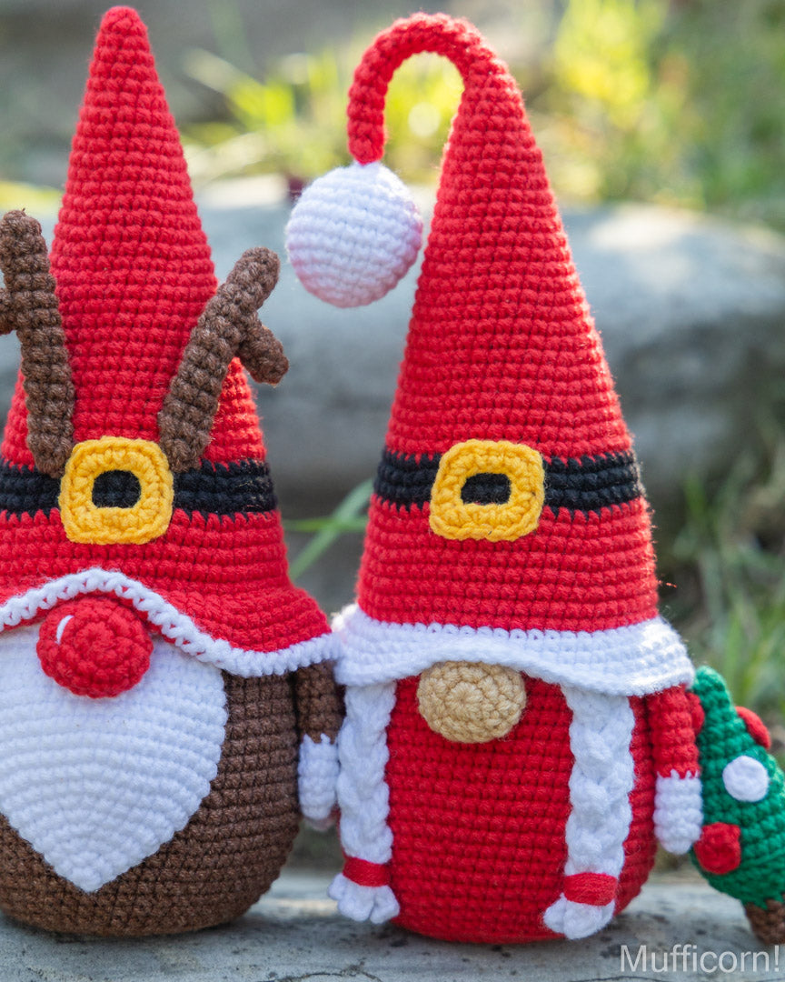 Two crocheted Christmas gnomes posed outdoors on a stone surface with a blurred natural background. One gnome has brown antlers and a white beard, while the other wears a red hat with a white pom-pom and a small green Christmas tree accessory.