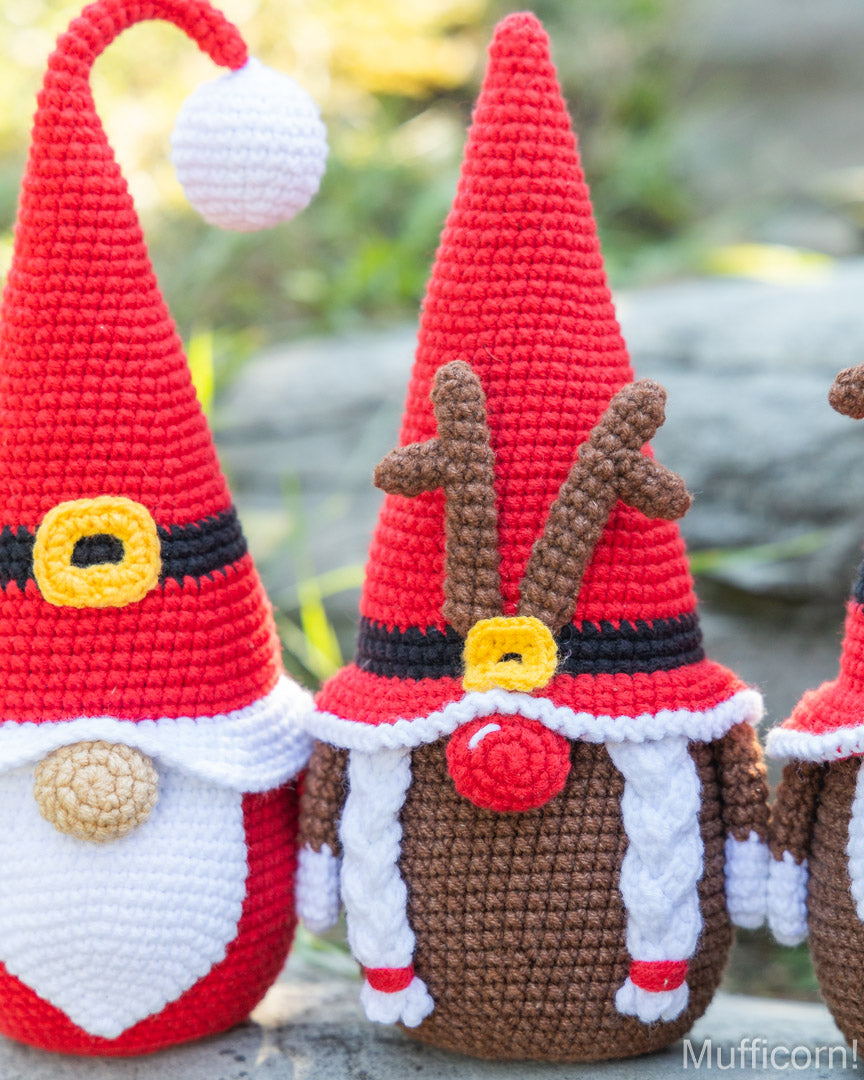 Handmade crocheted Christmas gnome and reindeer decorations, with one Santa Claus figure on the left and a reindeer on the right, shown outdoors against a blurred green background.
