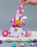 A crocheted Easter-themed decoration featuring a white bunny hat with pink ears, white daisy flowers, and a yellow bow, placed on top of a white crocheted pot filled with chocolate eggs. A hand is holding the hat, with pink and blue decorative eggs and foil chocolate candies scattered on a white surface against a gray background.